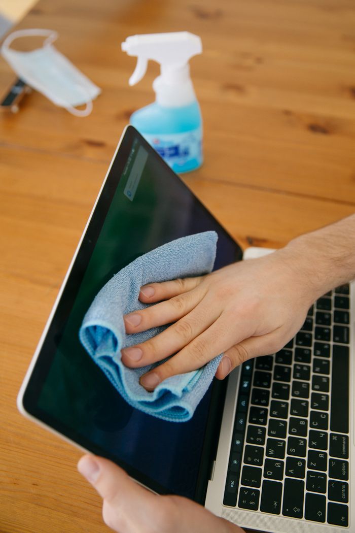creative-03 A person cleans a laptop screen with a blue cloth and spray bottle on a wooden table to keep it hygienic.